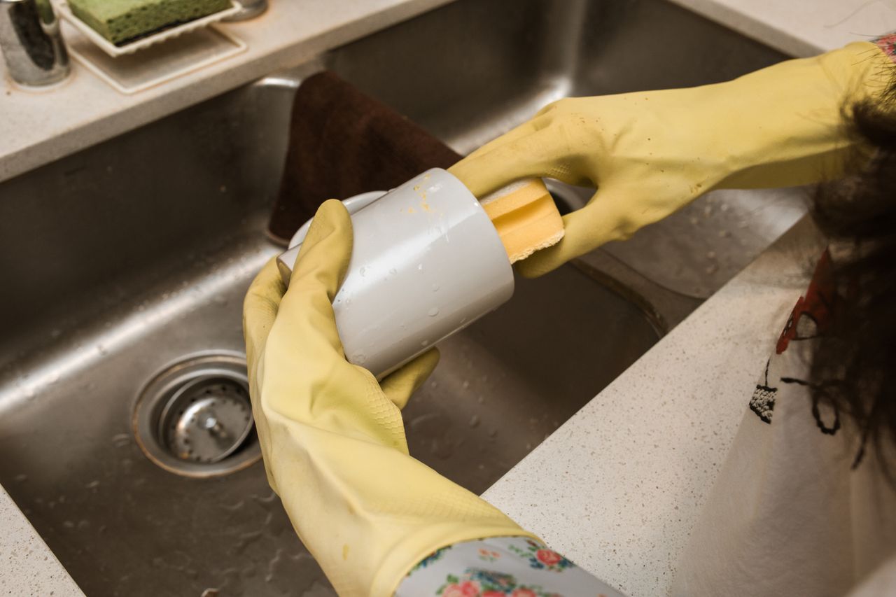journey-01 Close-up of a person washing a mug with yellow rubber gloves and sponge in a kitchen sink.