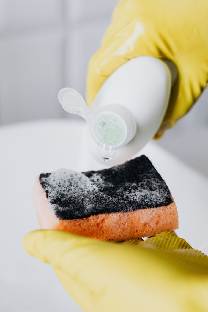 gallery-01 Unrecognizable person wearing yellow latex gloves holding orange sponge while pouring detergent in bathroom at modern home