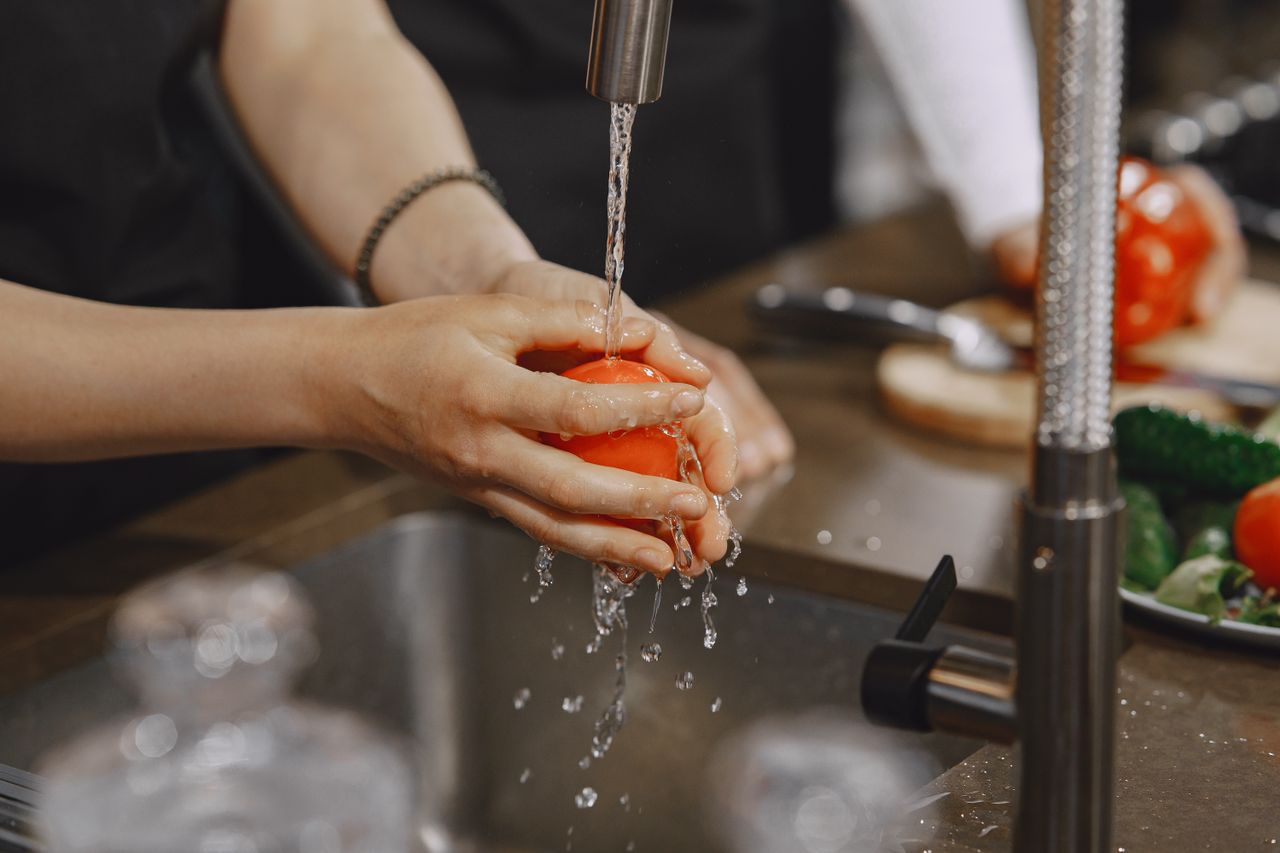 about-img-01 Close-up of hands washing a ripe red tomato under a kitchen faucet. Water splashing in action.