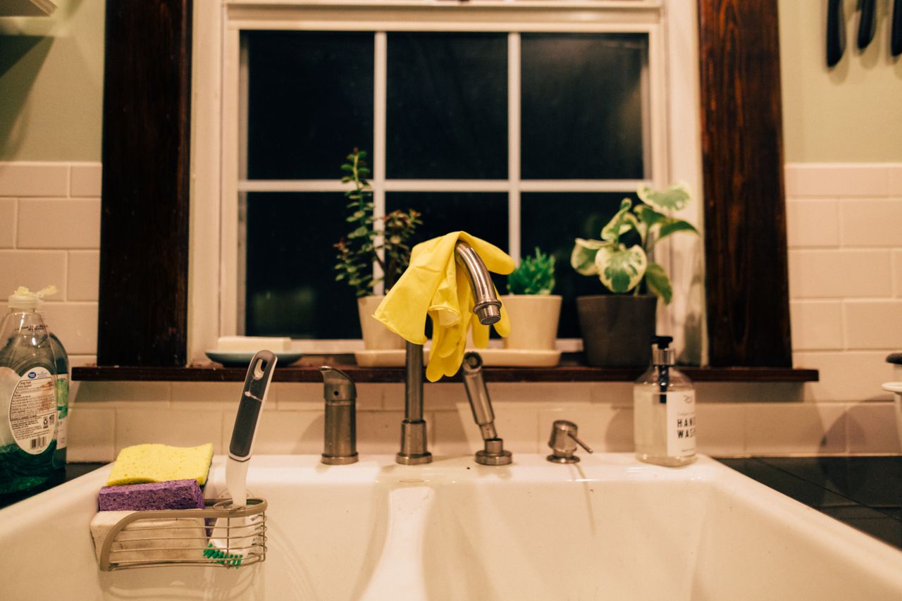 gallery-02 Warm lit kitchen sink at night with gloves, plants, and cleaning supplies.