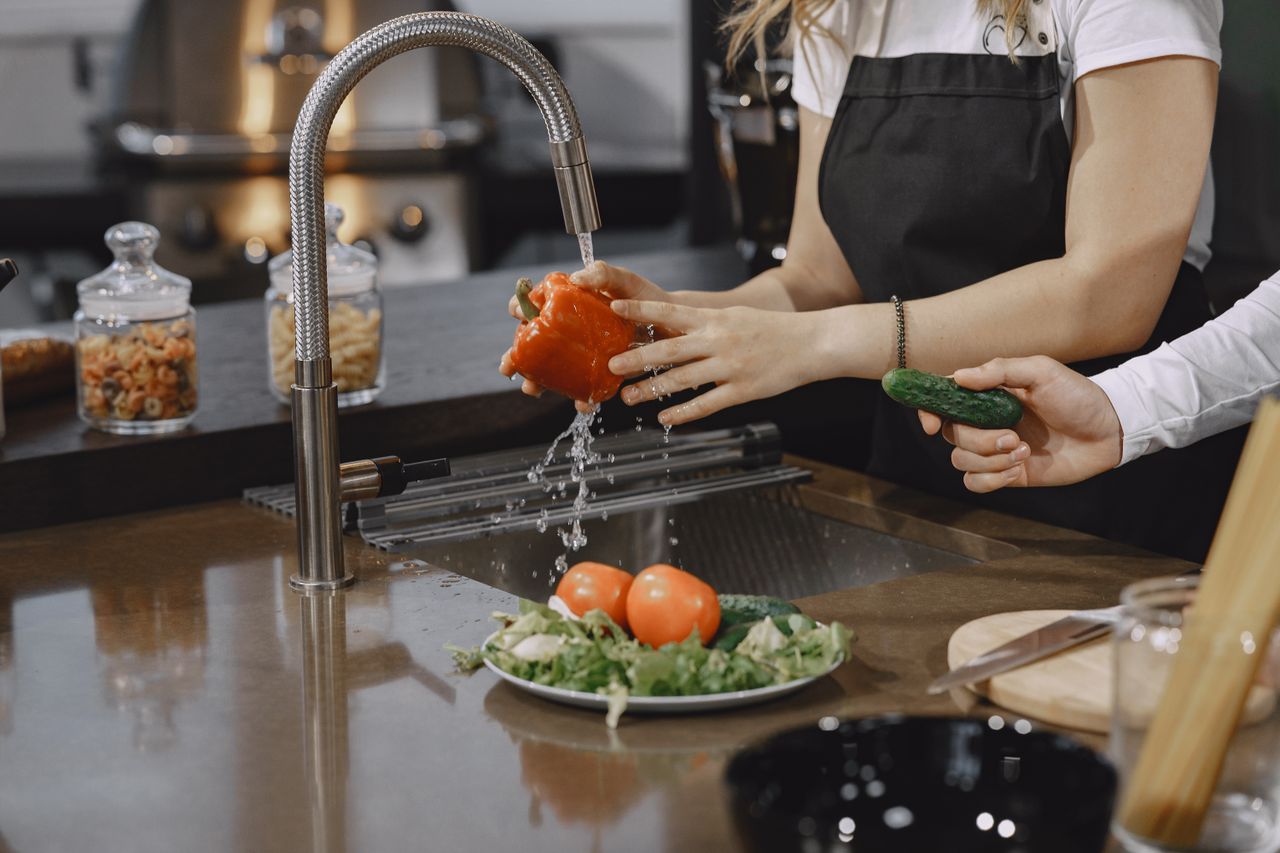 gallery-03 Hands washing a red bell pepper in a modern kitchen sink with fresh vegetables.